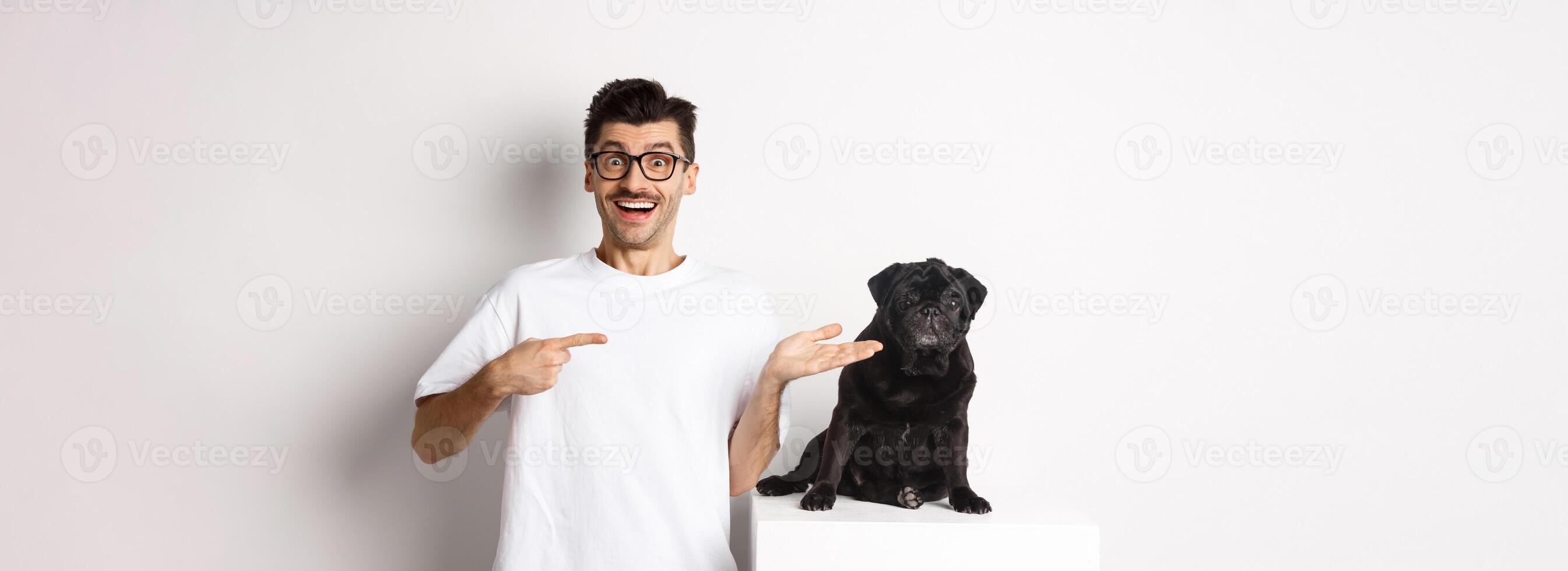 Cheerful young man pointing finger at his dog, showing small cute black pug sitting, white background photo