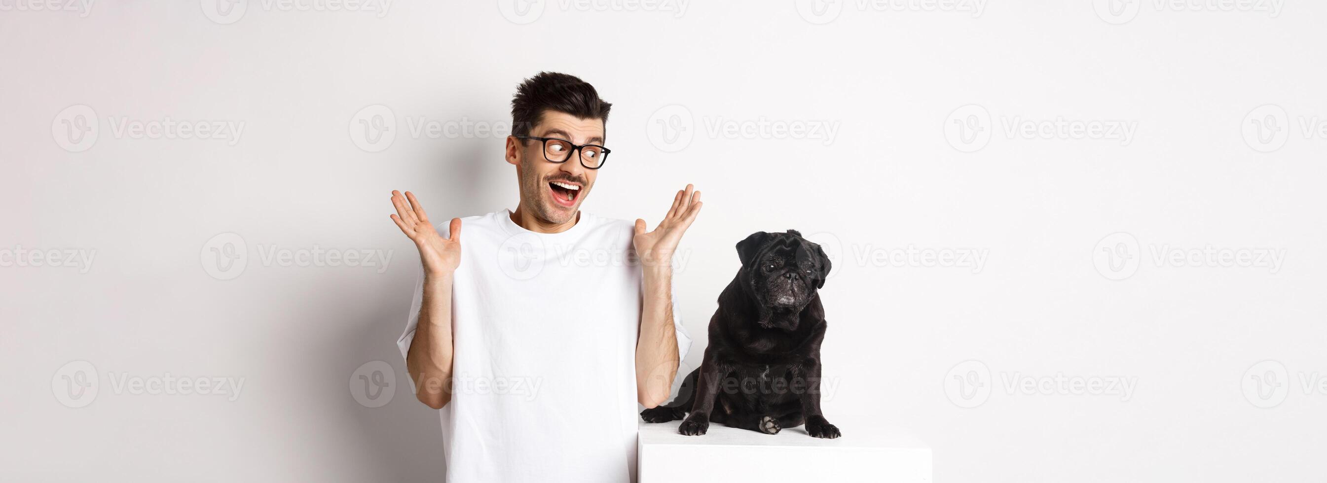 Image of cheerful young man rejoicing, looking at cute black dog pug and smiling, standing over white background photo