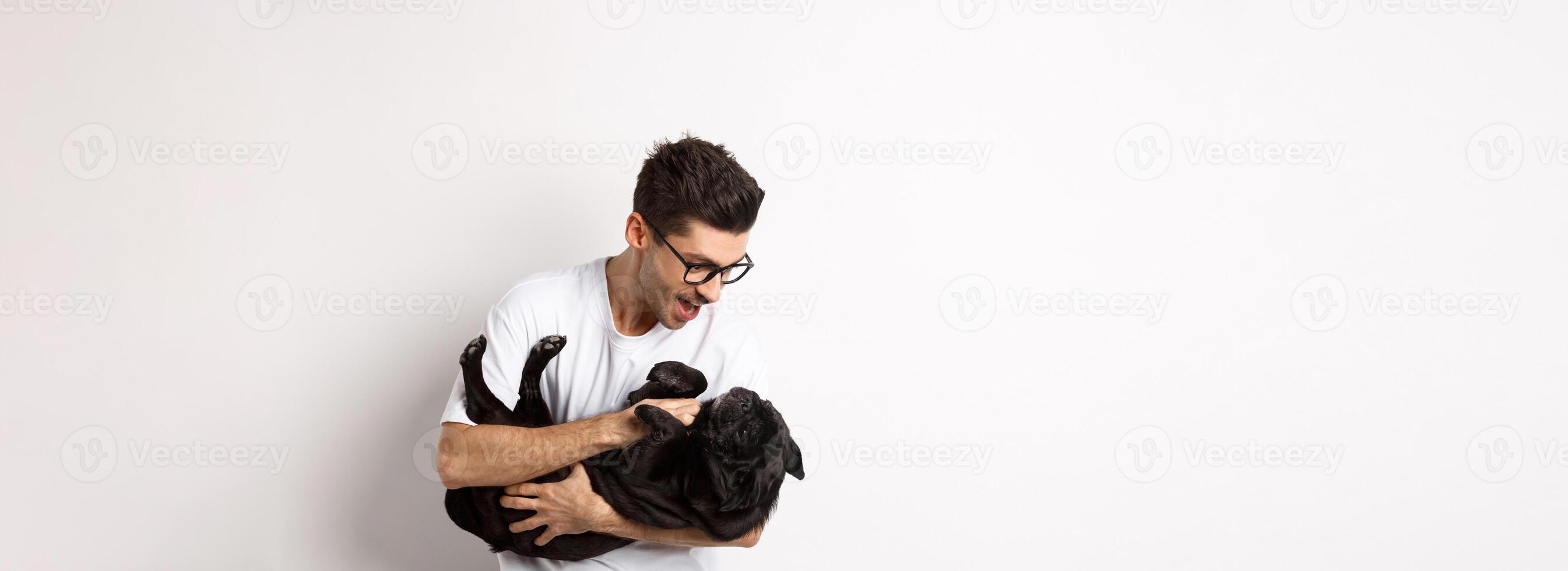 Handsome young man playing with cute black puppy. Dog owner petting a pug, standing over white background photo