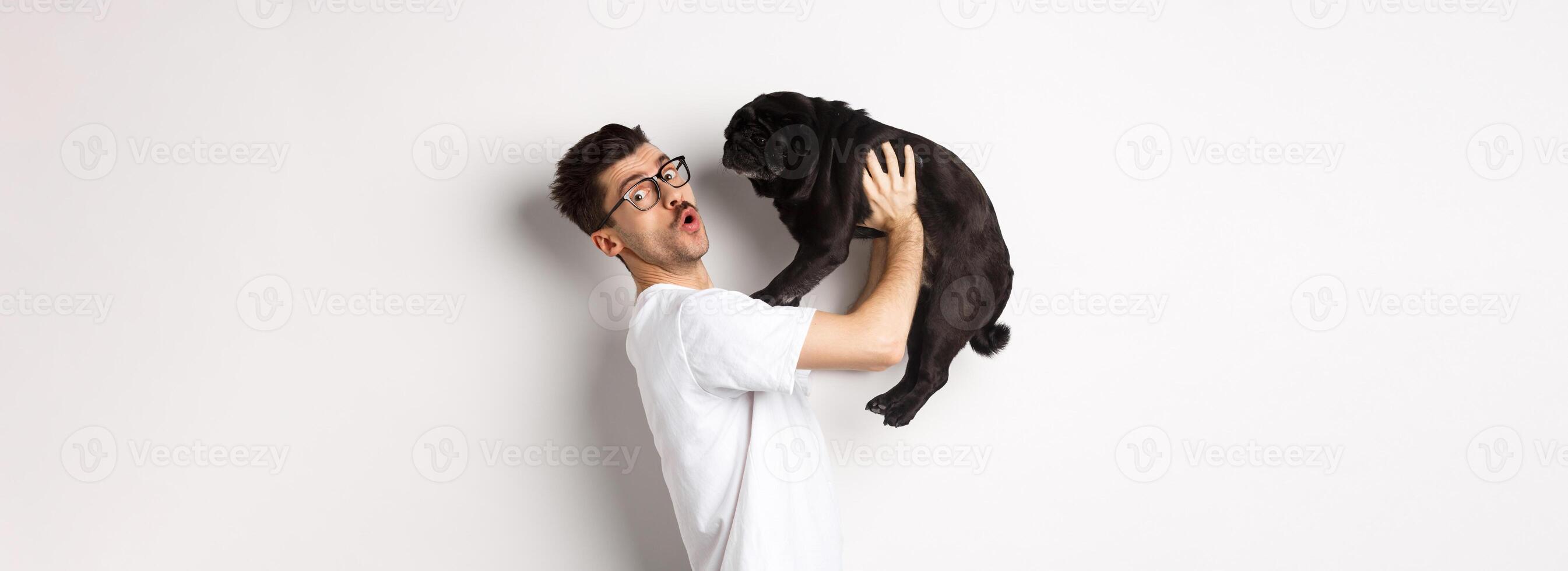 Image of handsome young man loving his pug. Dog owner holding puppy and smiling happy at camera, standing over white background photo