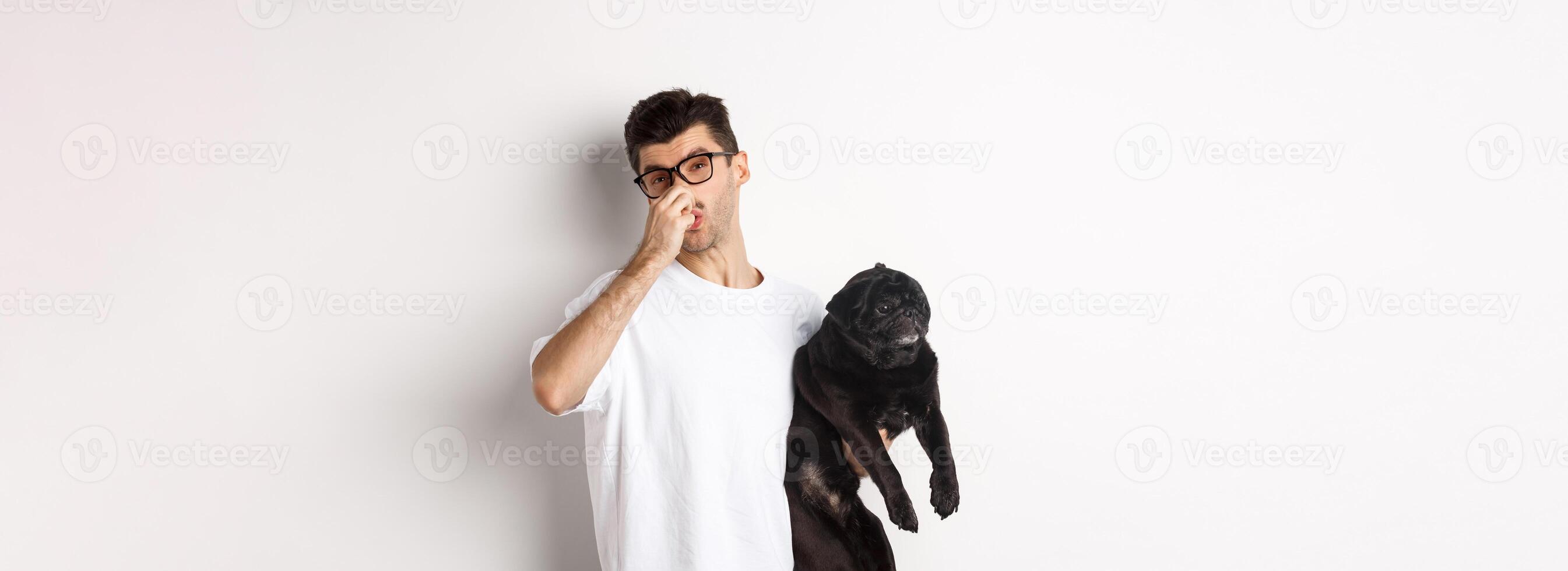 Young man shut nose as holding pug, disgusted with bad smell fart of animal, standing over white background photo