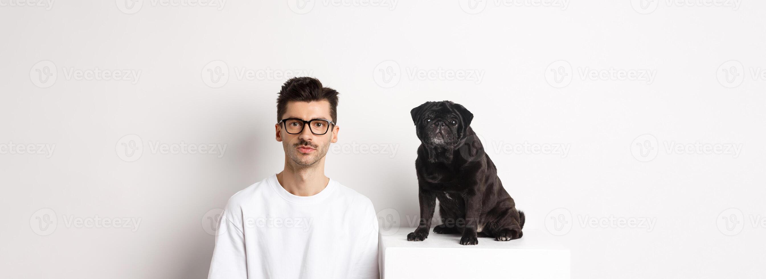 Image of handsome hipster in glasses sitting next to black cute pug dog, both staring at camera over white background photo