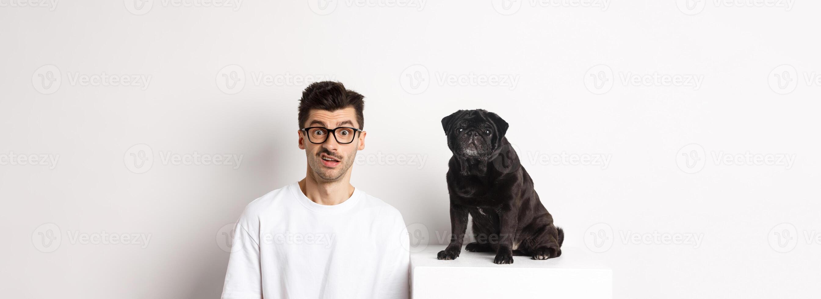 Image of hipster dog owner sitting near cute black pug, staring at camera confused and puzzled, white background photo