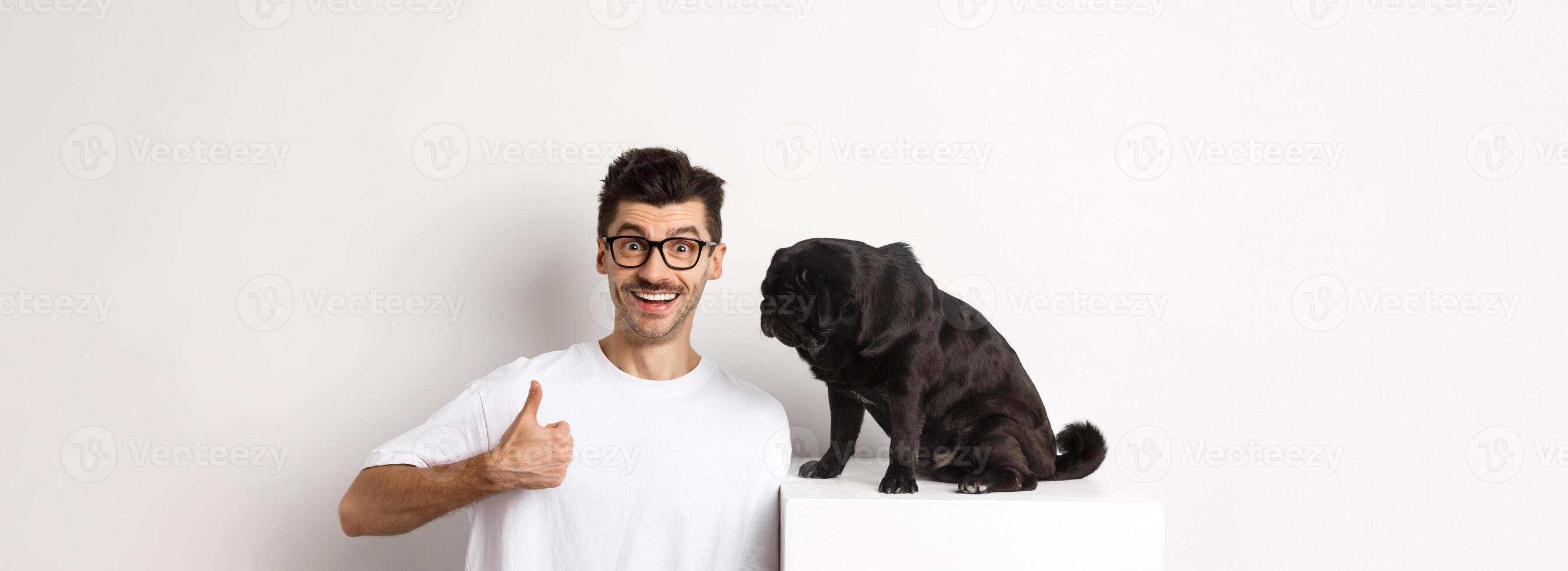 Image of happy and satisfied young man sitting near pug dog and showing thumbs-up, smiling and praising good product, white background photo