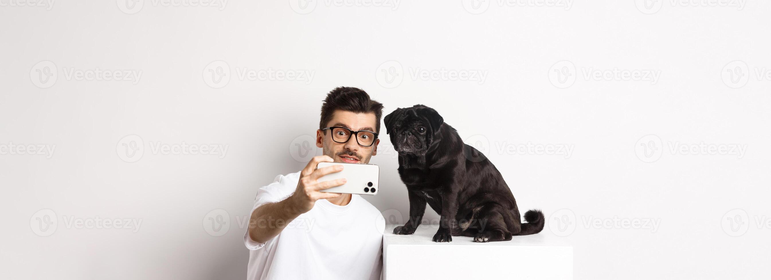 Image of handsome young man taking selfie with cute black dog on smartphone, posing with pug over white background photo