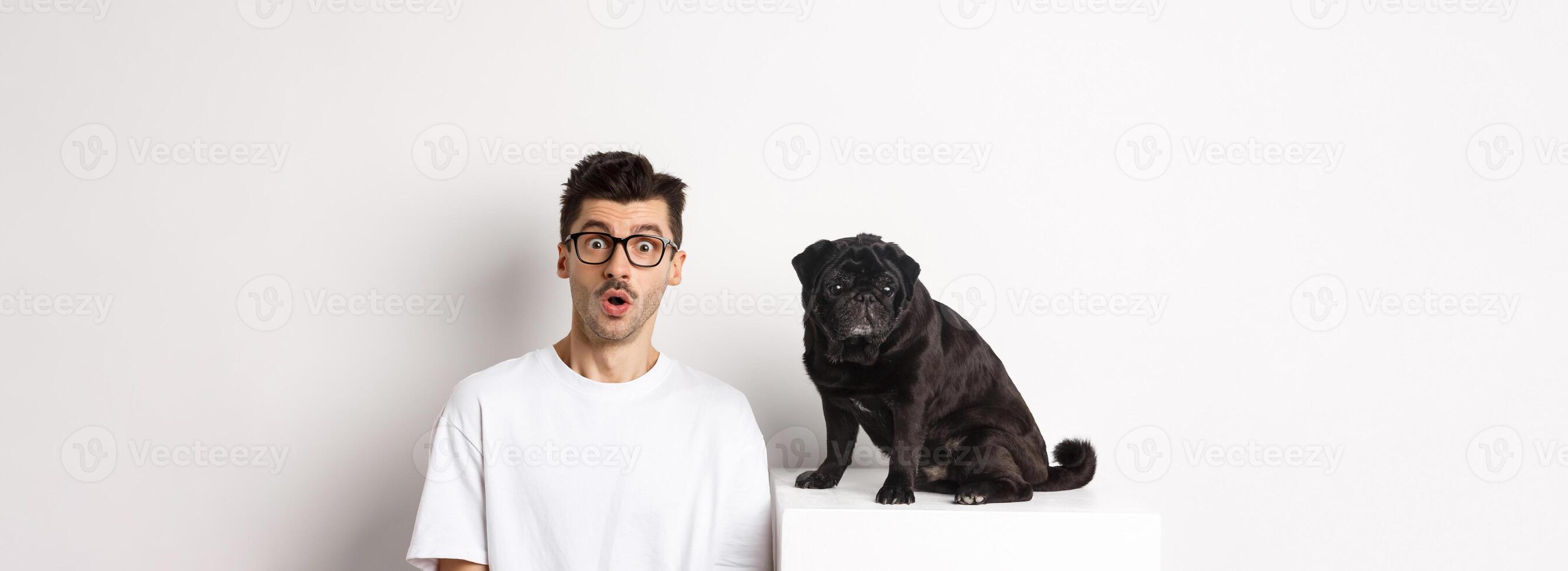 Portrait of dog owner and small cute pug staring at camera surprised and amazed, standing over white background photo