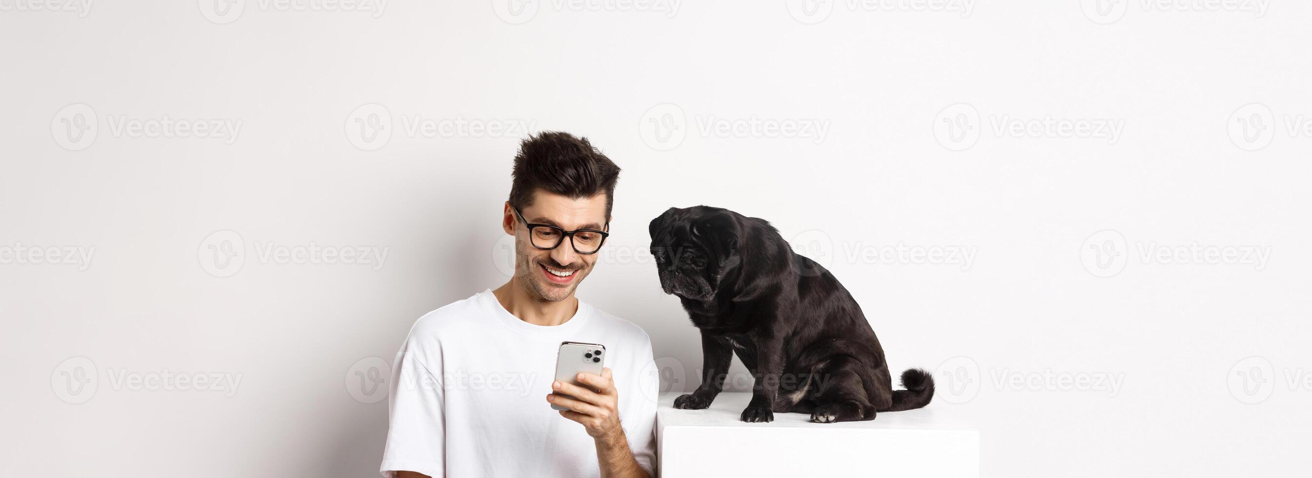 Smiling young man using smartphone and sitting near dog. Pug owner checking photos on mobile phone, white background