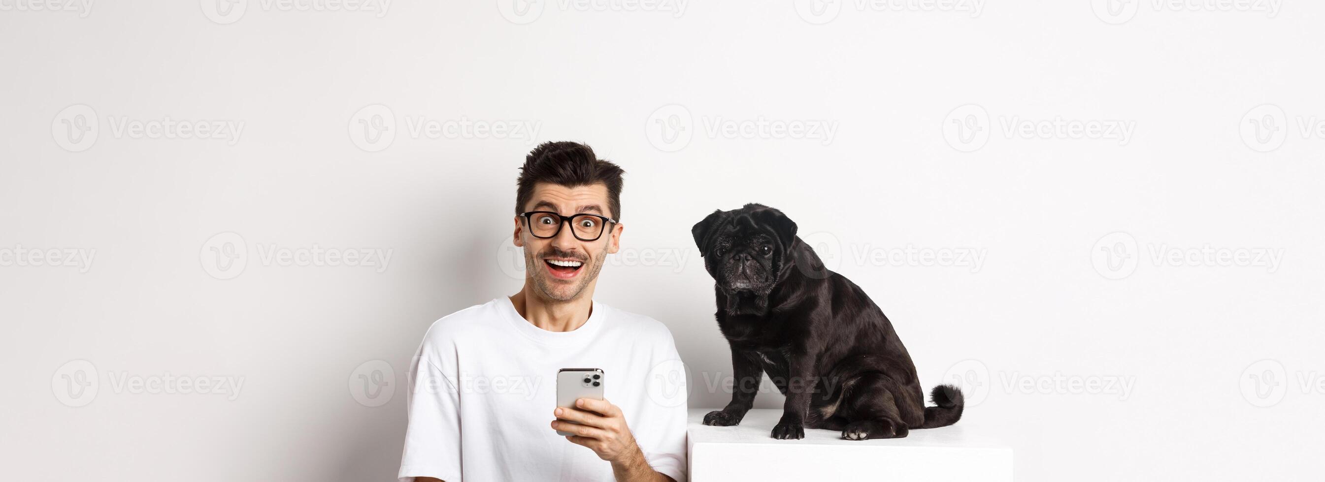 Cheerful young man hipster staring at camera, sitting with cute black pug dog and using mobile phone, standing over white background photo