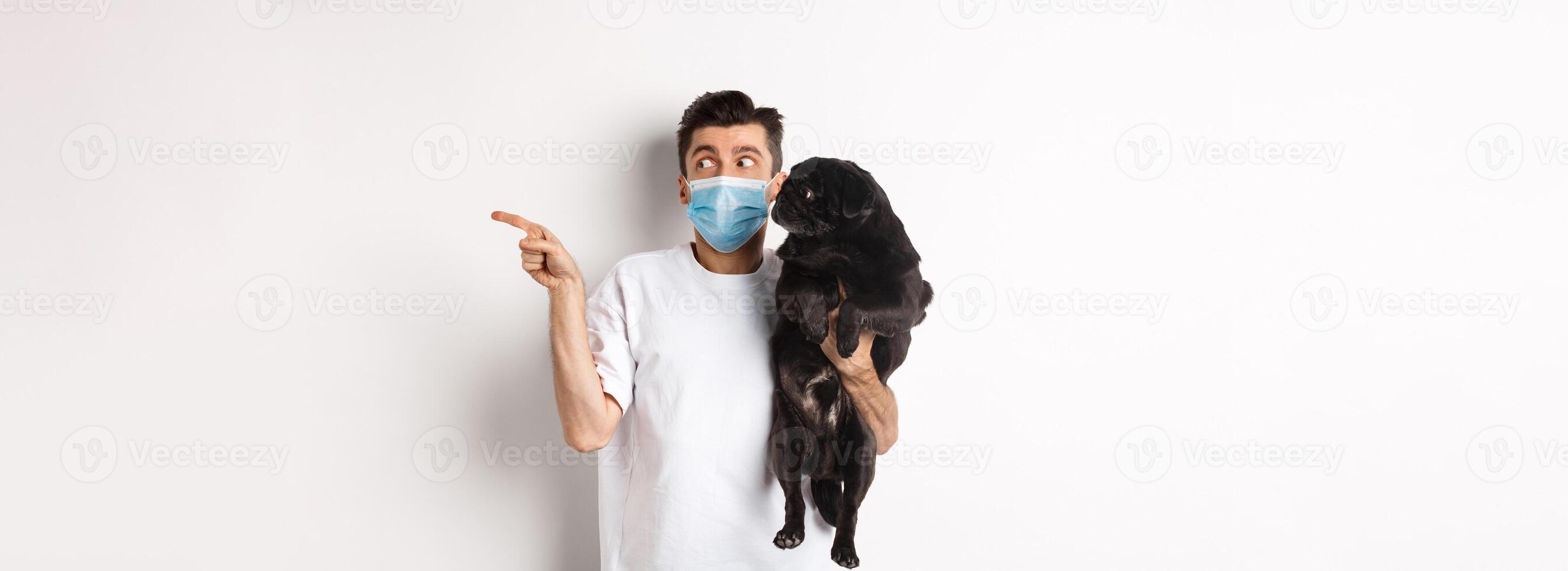 Covid-19, animals and quarantine concept. Young man in face mask holding cute black pug, dog and pet owner looking right at copy space, standing over white background photo