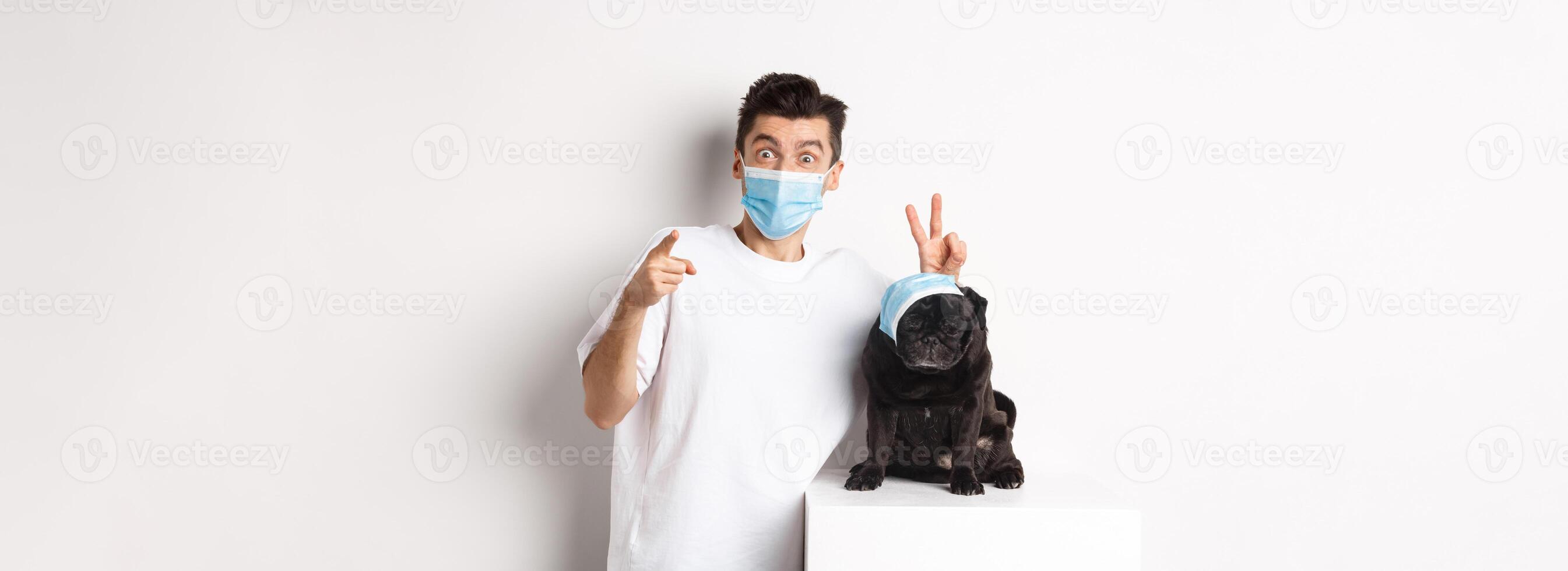 Covid-19, animals and quarantine concept. Happy dog owner and cute pug wearing medical masks, man pointing finger at camera and making funny bunny ears on pet, white background photo