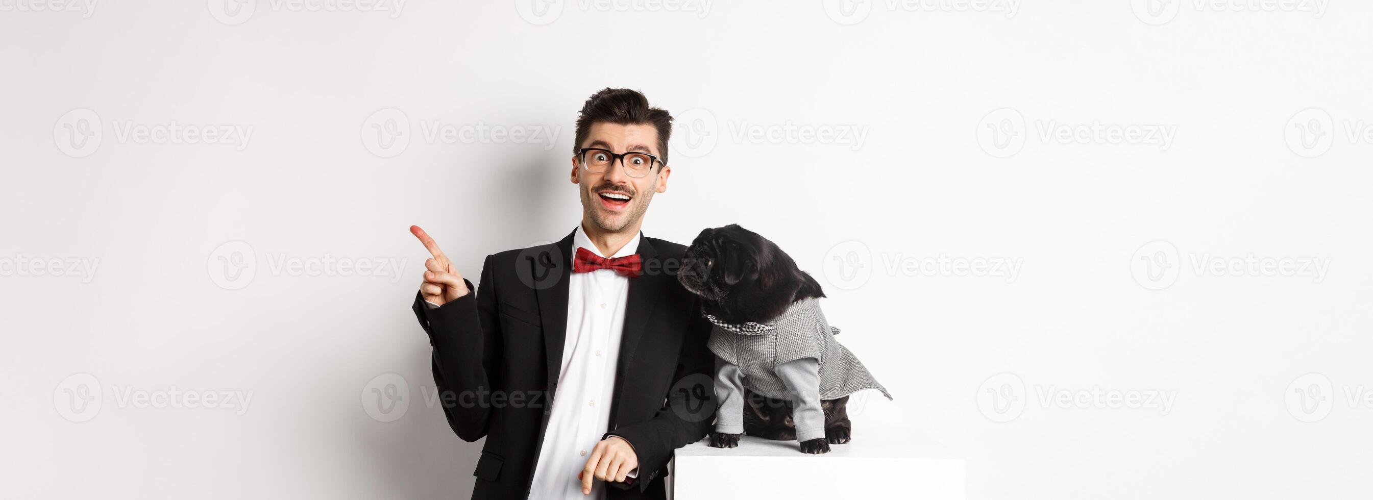 Animals, party and celebration concept. Happy and amazed young man and cute pug in costume standing over white background, dog owner pointing right photo