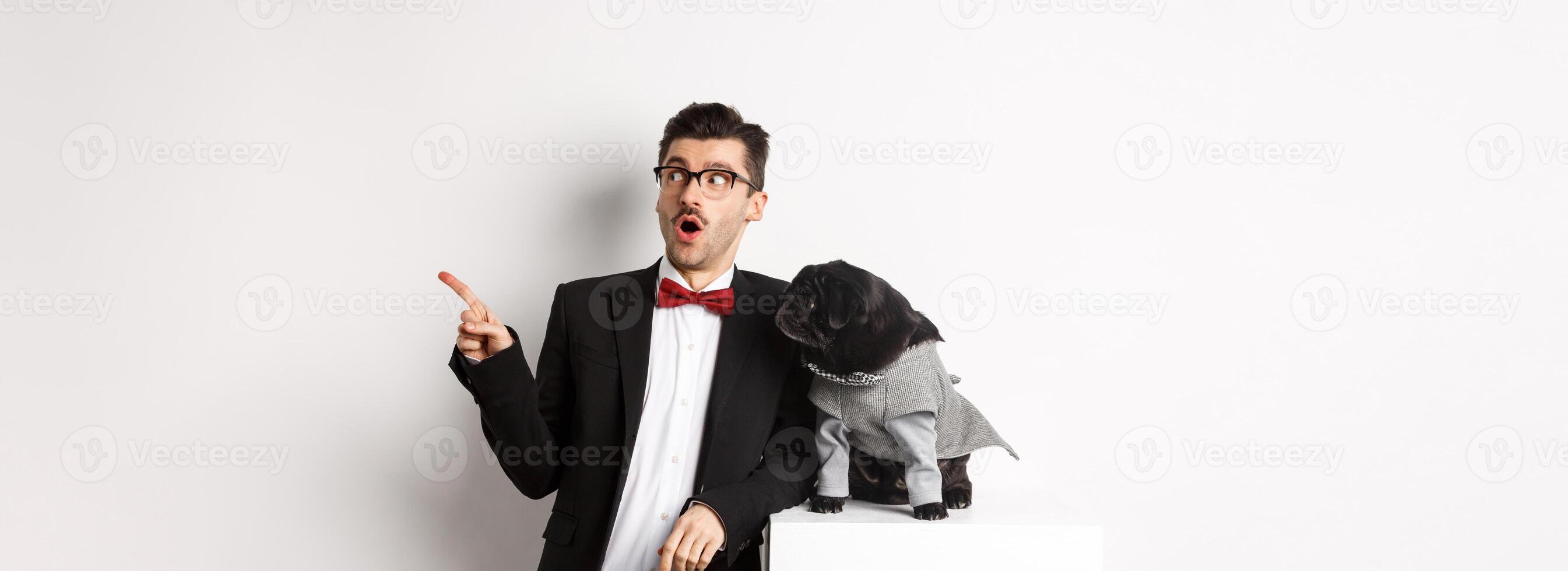 Animals, party and celebration concept. Amazed young man and black dog in costumes staring right at copy space, standing against white background photo