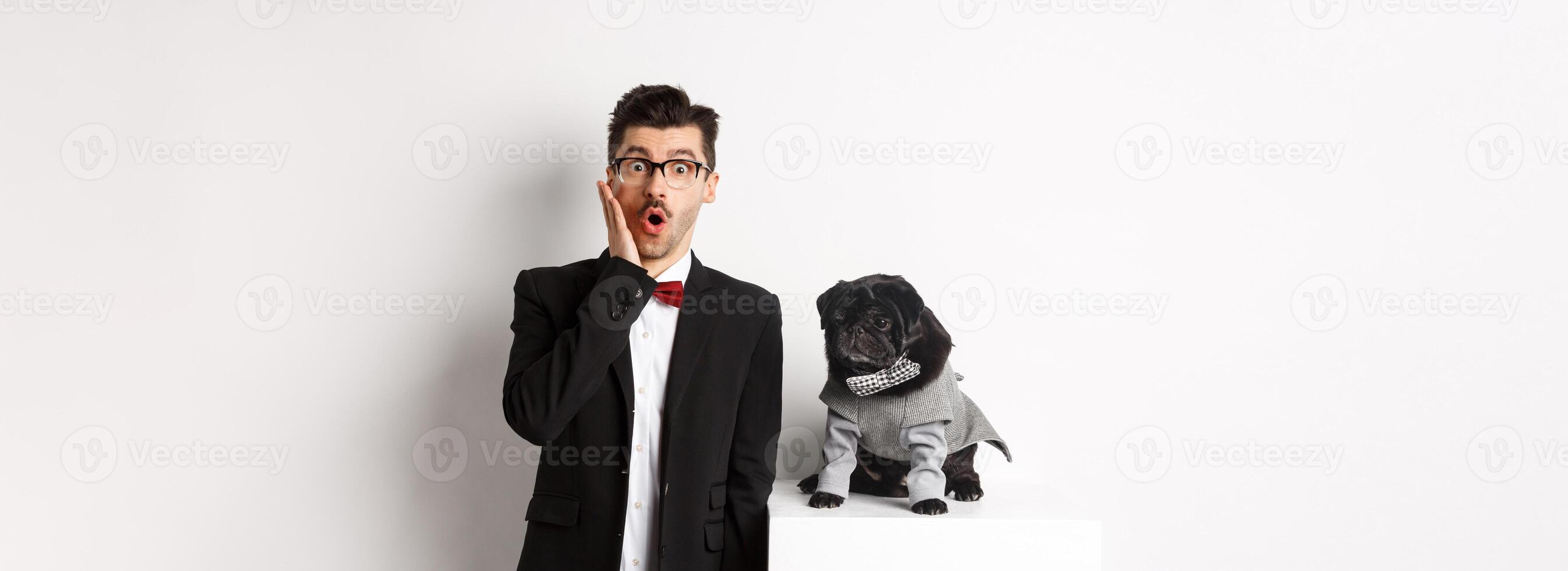 Animals, party and celebration concept. Shocked handsome man in formal suit and cute dog in costume, staring at camera amazed, standing over white background photo
