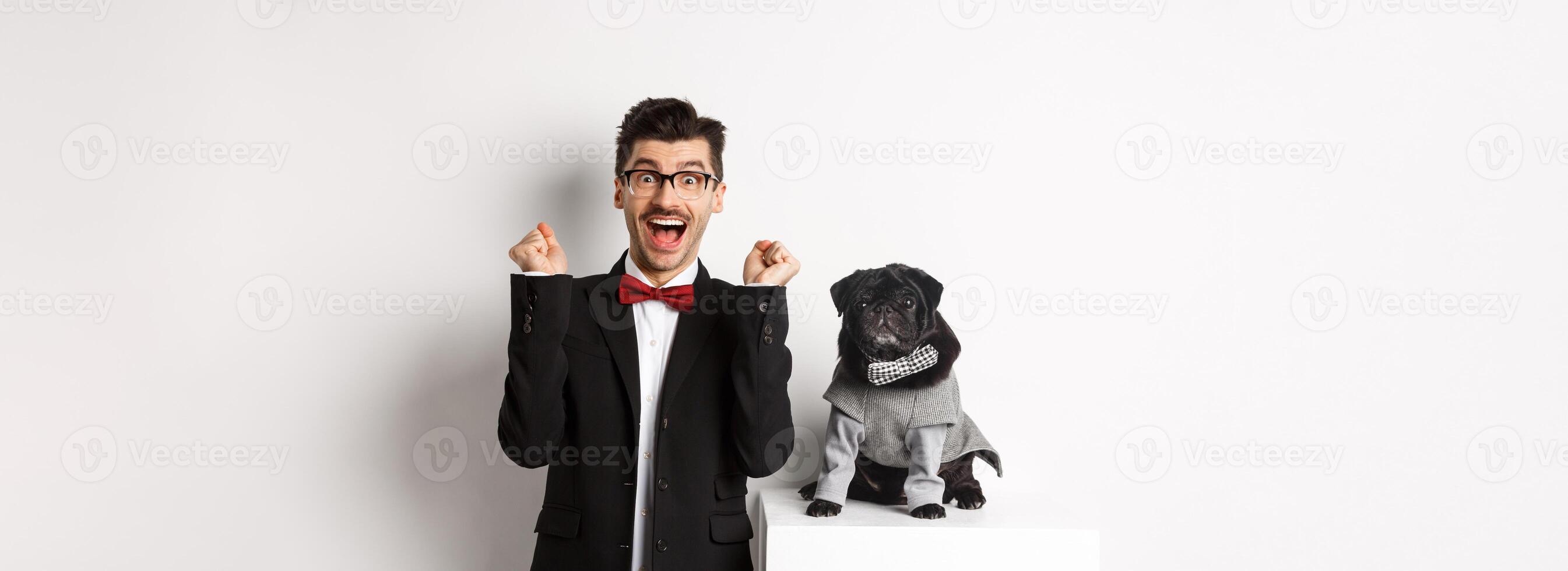 Animals, party and celebration concept. Happy young man in suit and puppy in pet cosume standing over white background, dog owner rejoicing and triumphing photo