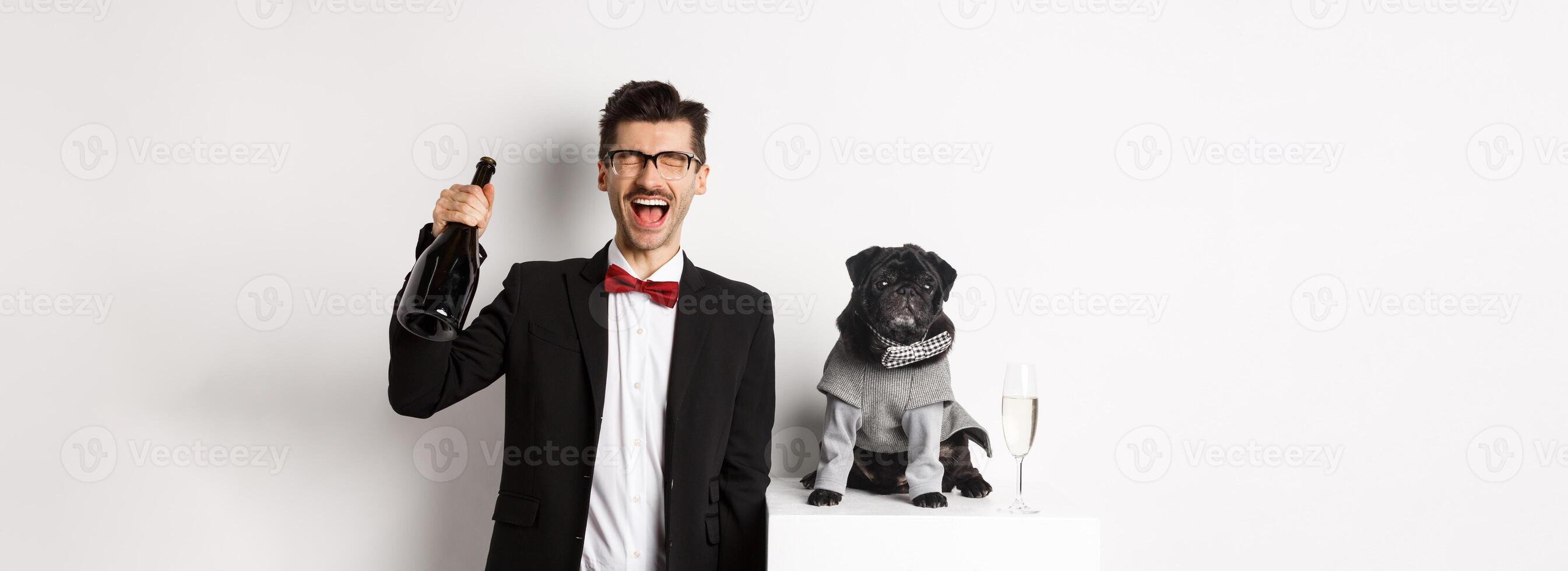 Pets, winter holidays and New Year concept. Happy man celebrating Christmas party pet, standing with cute dog in costume, drinking champagne and rejoicing, white background photo