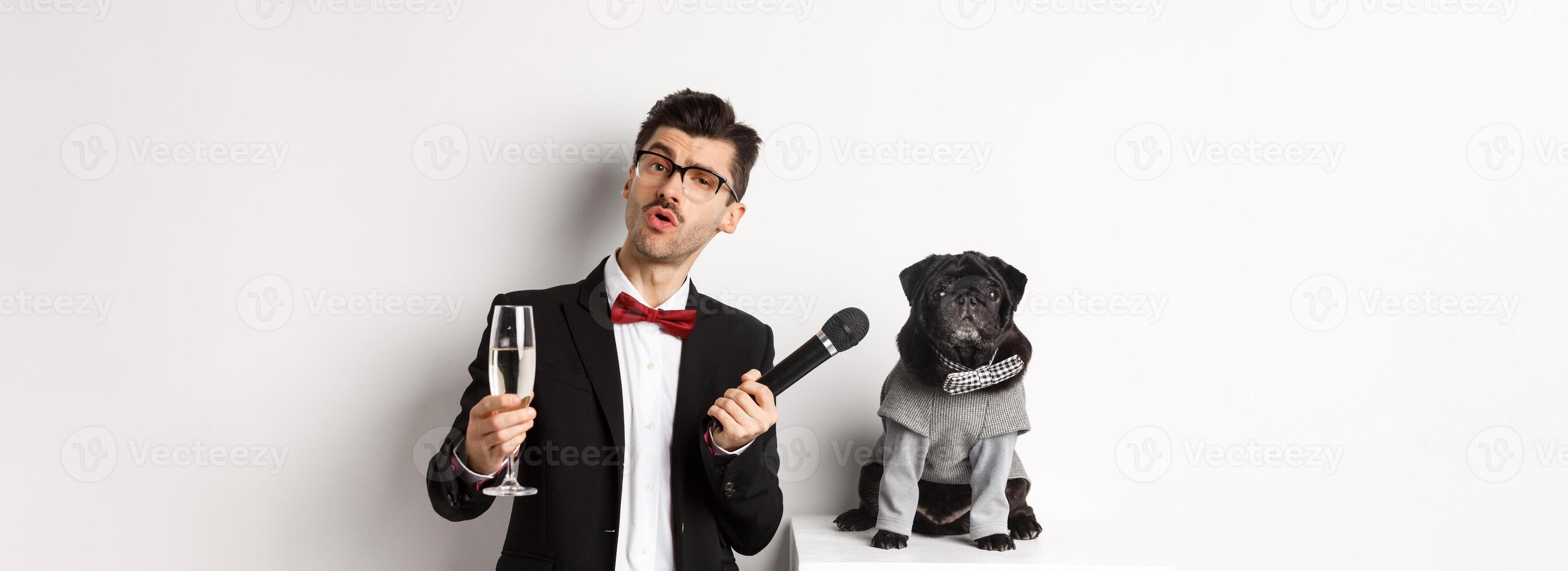 Handsome fancy man in glasses, raising glass of champagne and giving microphone to cute pug in party suit, celebrating and having fun, white background photo
