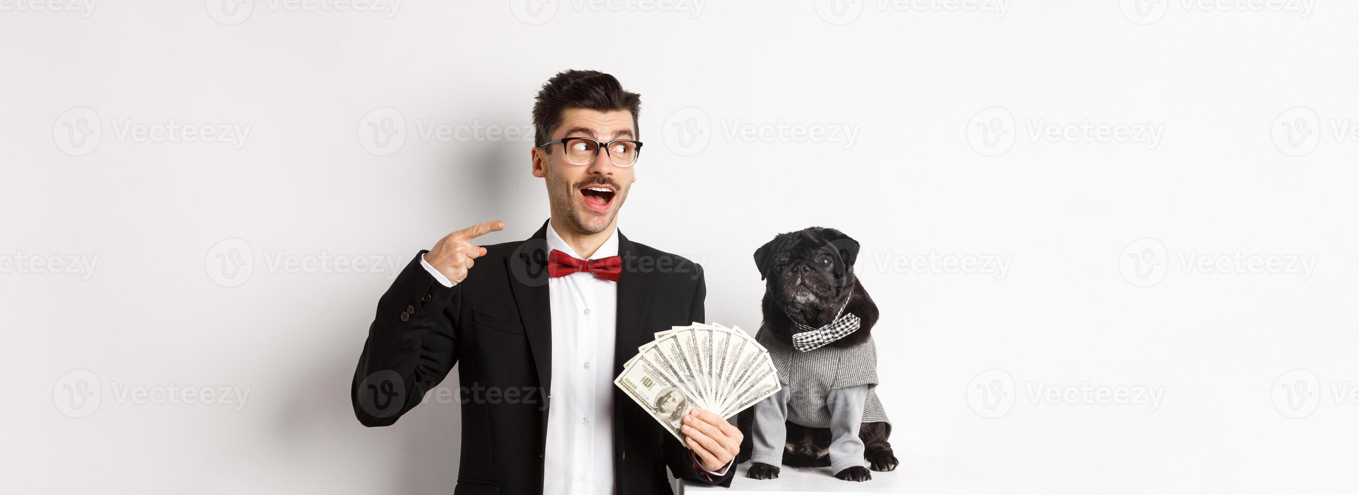 Happy young man in suit earn money with his dog. Guy rejoicing, holding dollars and pointing left, black pug in costume staring at camera, white background photo