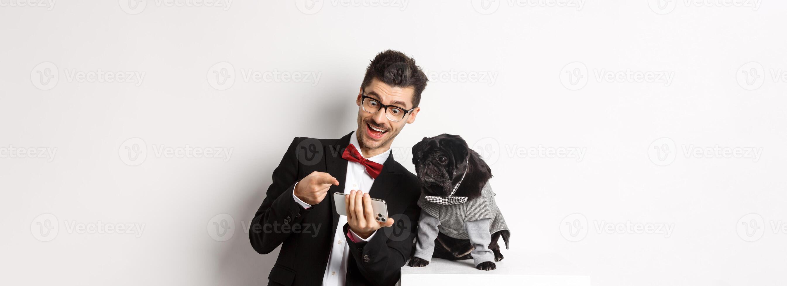 Happy dog owner showing something to pet on mobile phone, man and pug wearing fancy costumes, standing over white background photo