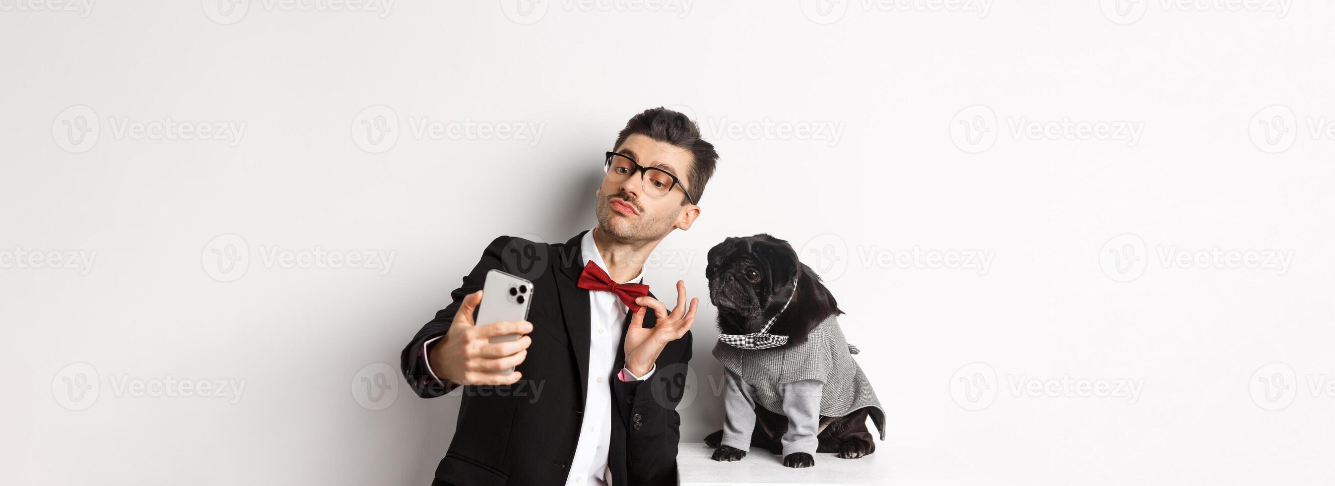 Cheerful dog owner in suit celebrating New Year with dog, taking selfie on smartphone near cute black pug in costume, white background photo