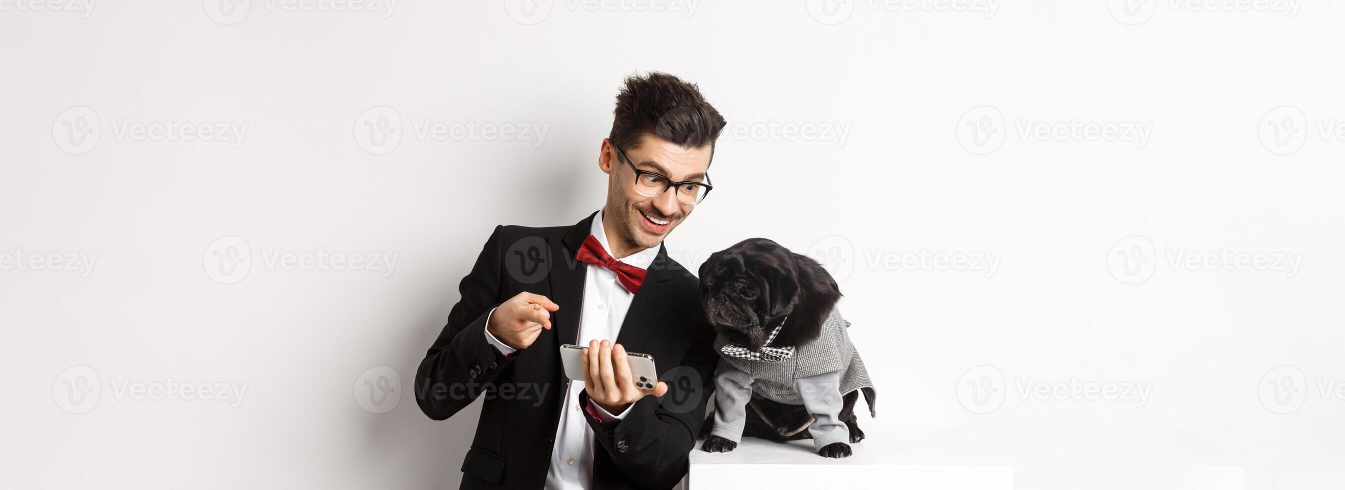 Handsome young man showing something on mobile phone to his dog. Owner shopping online with pet, standing in costumes over white background photo
