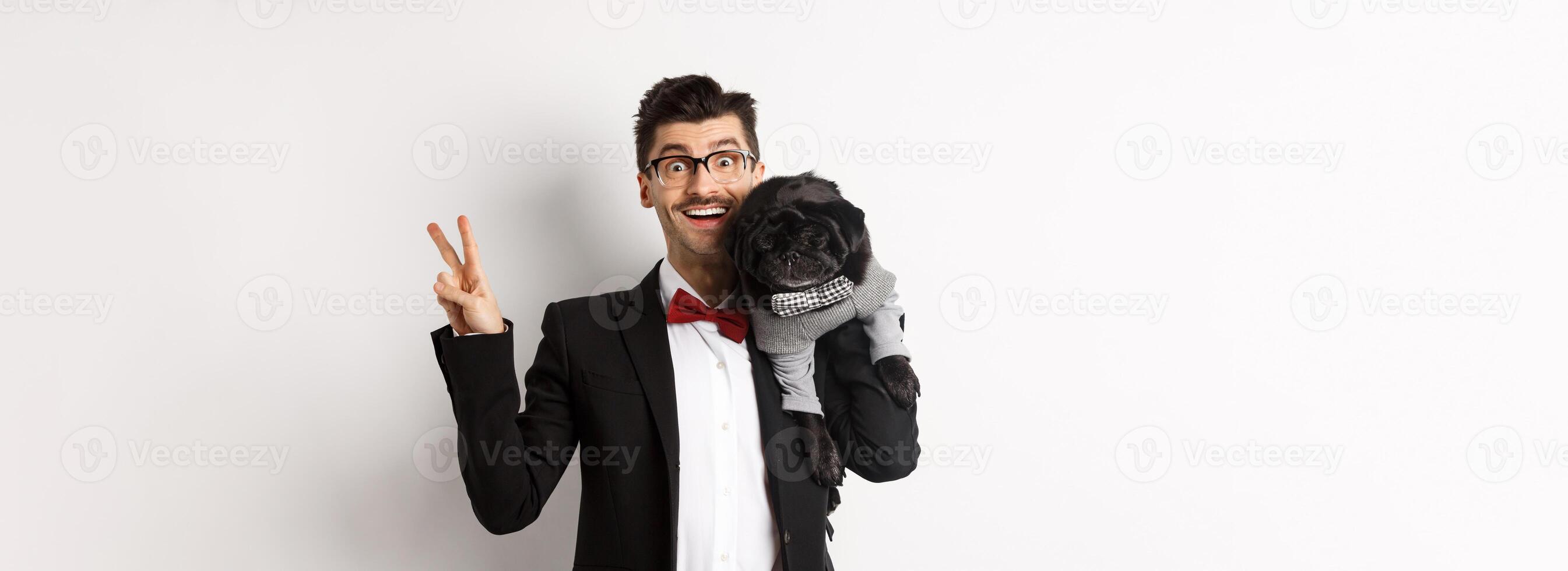 Cheerful young man in suit and glasses taking photo with cute black pug dog on his shoulder, smiling happy and showing peace sign, posing over white background