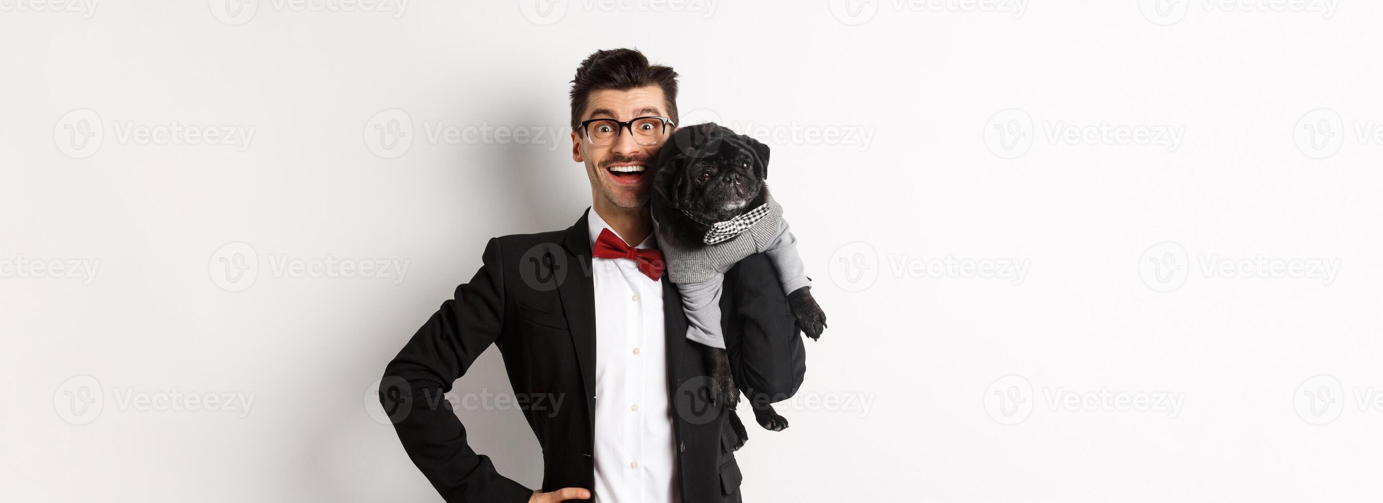 Handsome young man in suit and glasses holding cute black pug dog on shoulder, smiling happy at camera, wearing party outfits, white background photo