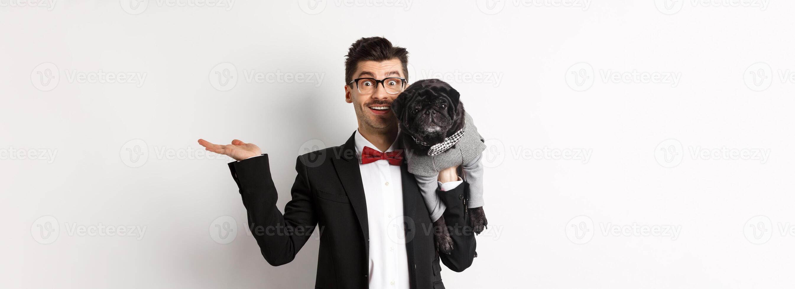 Handsome young man introducing pet product on hand, holding cute black dog on shoulder and smiling, showing something on white background photo