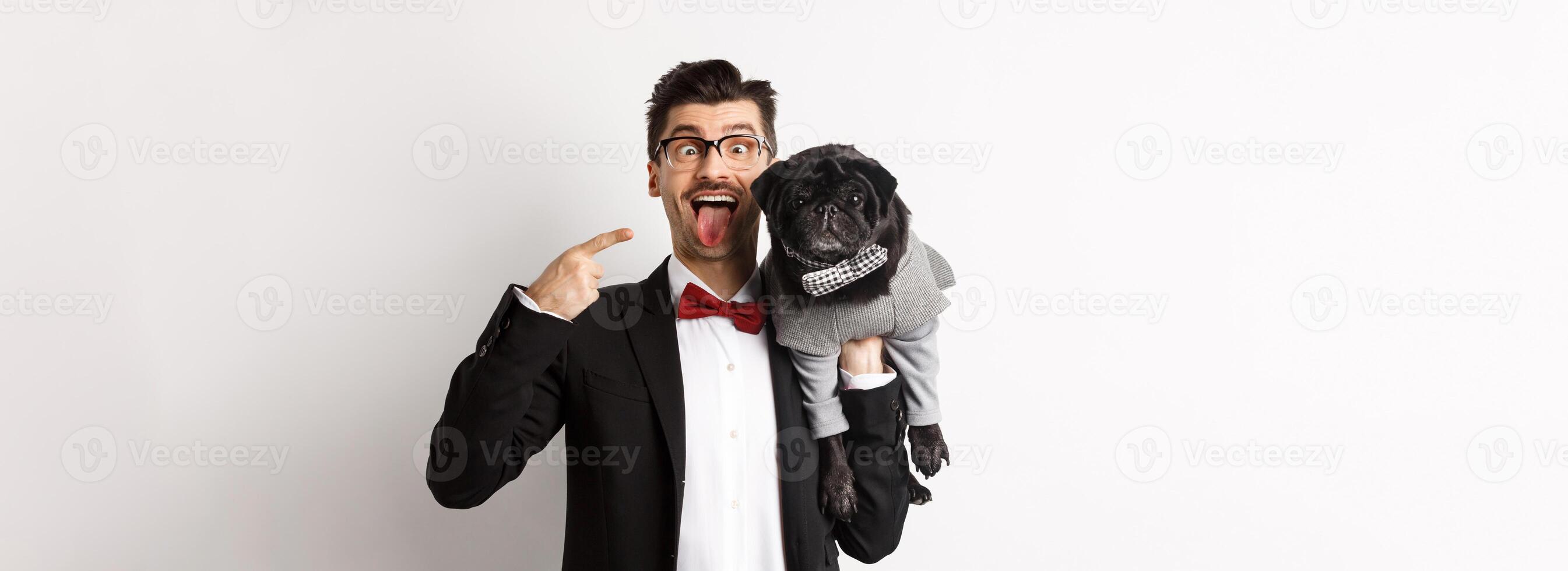 Funny young man in party suit, showing tongue and making grimaces, pointing at cute black dog in winter clothes, standing over white background photo