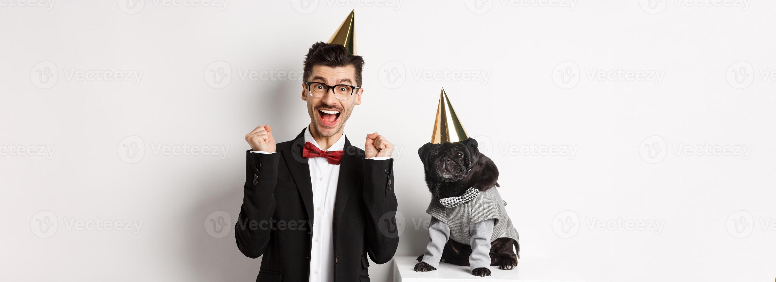 Cheerful young man shouting for joy, dog and owner wearing birthday party cones and celebrating, guy rejoicing and staring at camera, white background photo