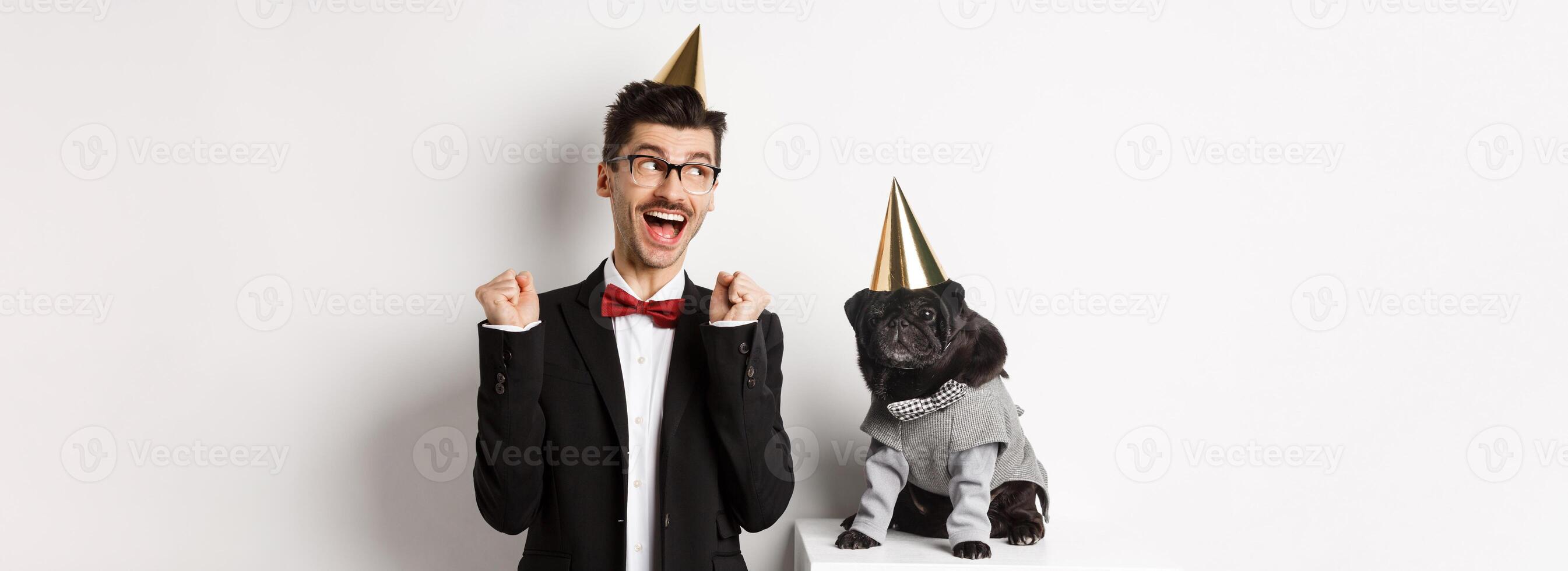 Cheerful young man shouting for joy, dog and owner wearing birthday party cones and celebrating, guy rejoicing and staring at camera, white background photo