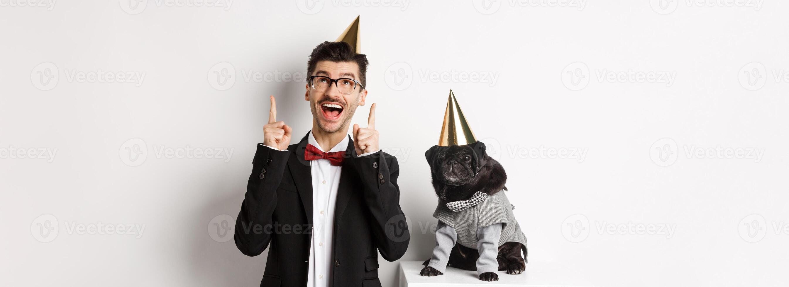 Cute black pug dog wearing party cone and standing near happy owner, man pointing fingers up at copy space, celebrating birthday, white background photo