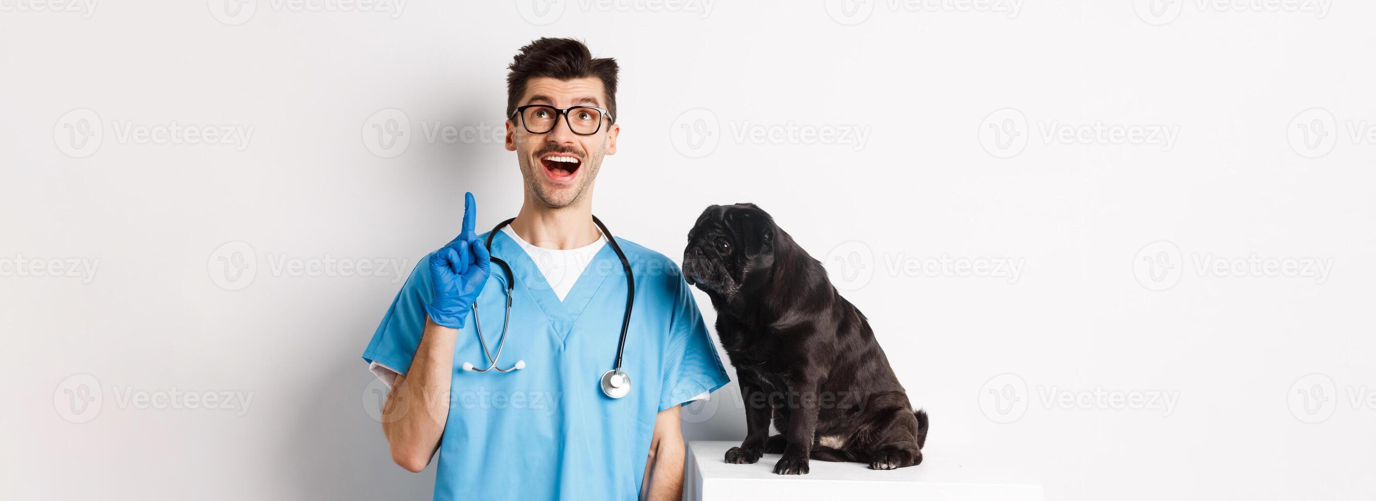 Handsome young doctor at vet clinic pointing finger up and looking amazed, standing near cute black pug dog, white background photo