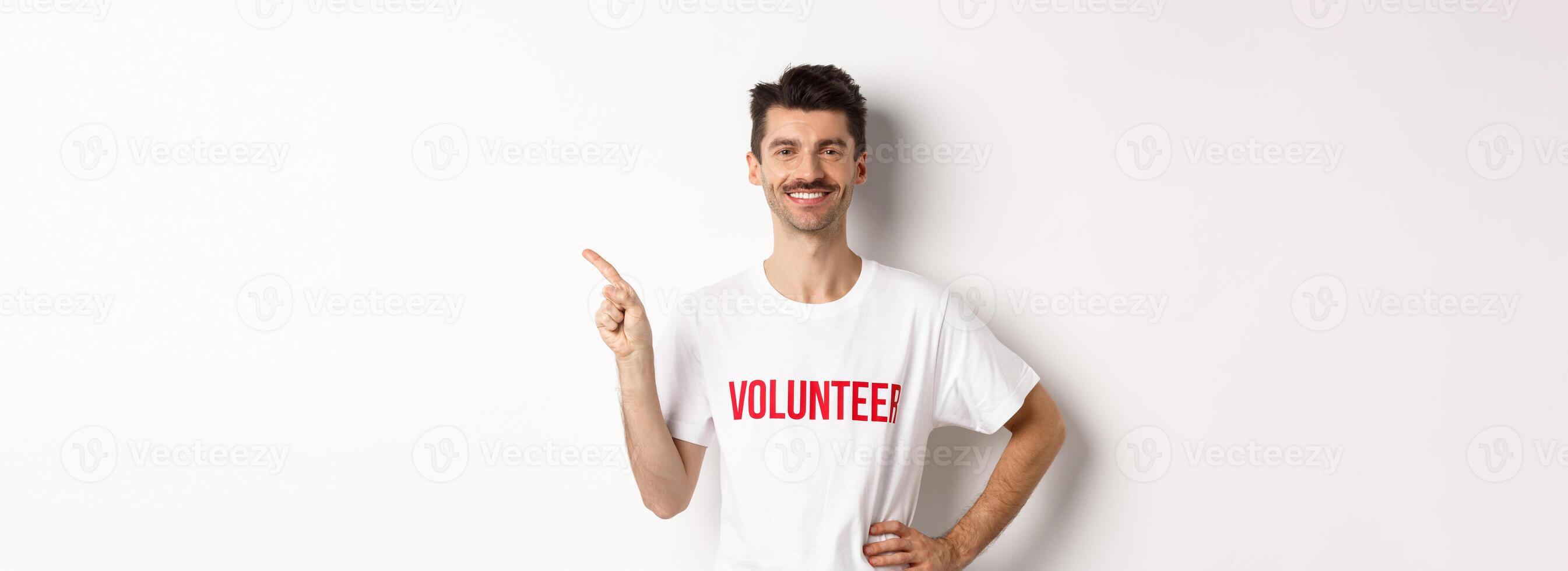 Handsome smiling volunteer in t-shirt pointing finger left, showing announcement, standing over white background photo