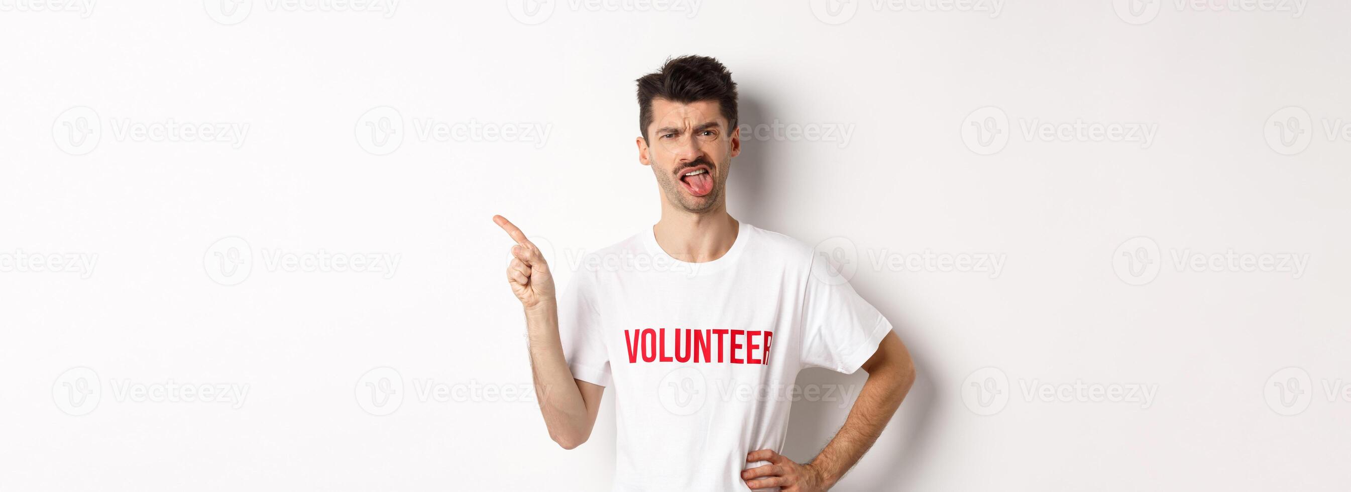 Disappointed volunteer showing bad promo offer, sticking tongue and grimacing disgusted, pointing finger left, wearing organization t-shirt, white background photo