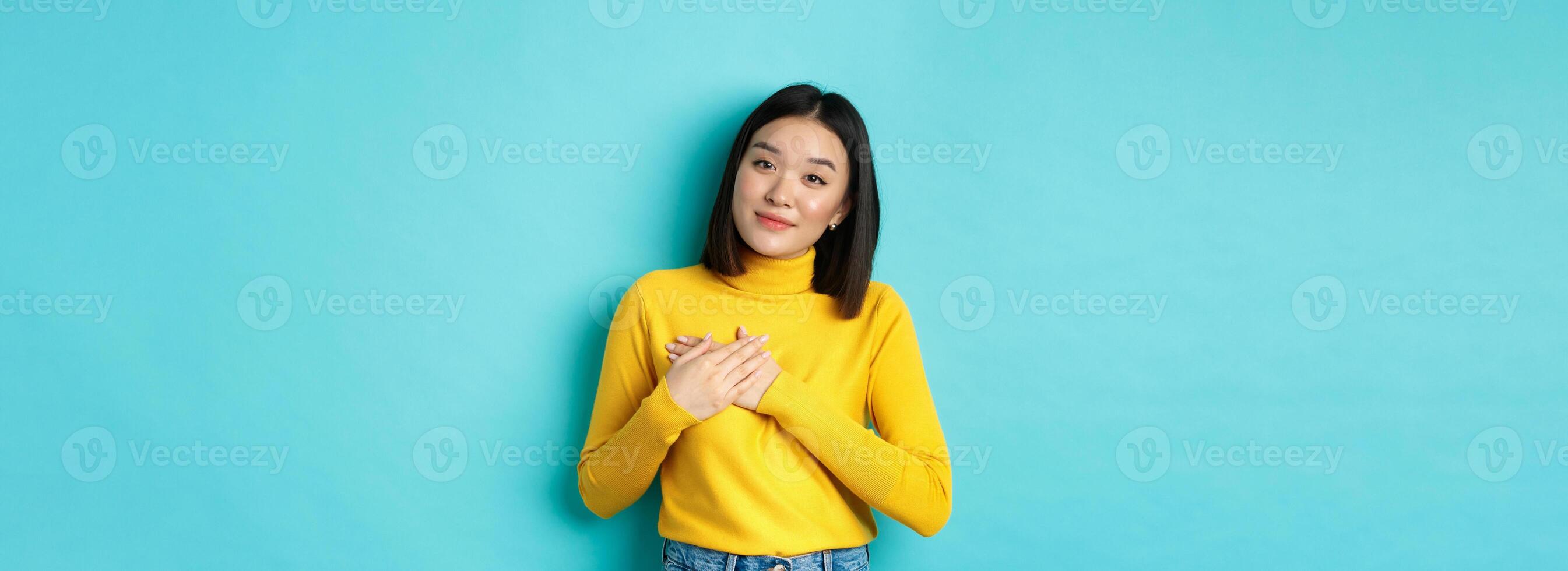 Portrait of beautiful heartfelt woman holding hands on heart, smiling and listening compassionate, standing over blue background in yellow pullover photo