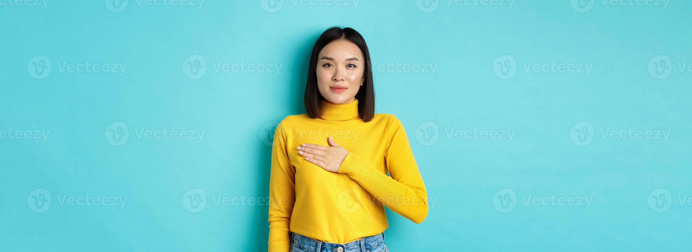 Image of proud smiling asian woman holding hand on heart, showing respect to national anthem, standing over blue background photo