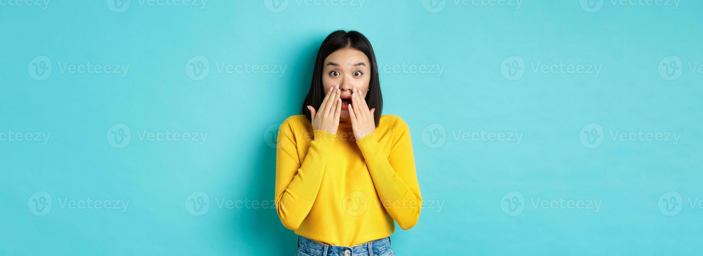 Portrait of impressed korean girl saying wow, gasping and staring amazed at camera, standing in yellow pullover over blue background photo