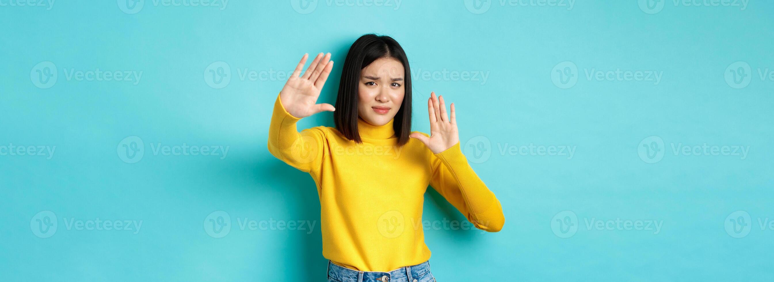 Image of woman feeling uncomfortable in front of camera, cover face with hands and frowning, begging to stop, standing against blue background photo