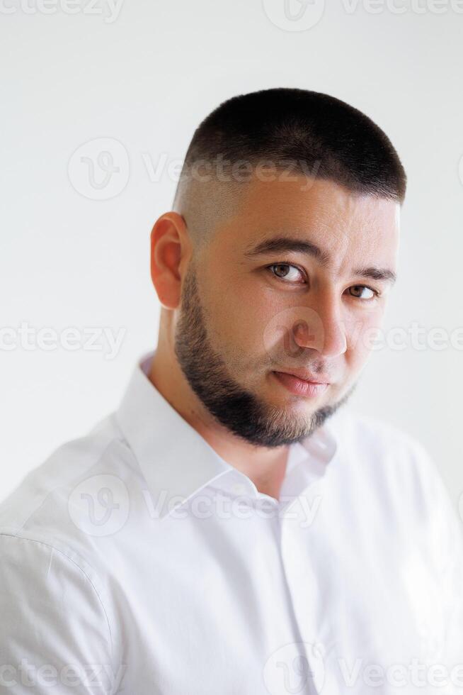 A man with a shaved head and a beard is standing in front of a white wall. He is wearing a white shirt and a white tie photo