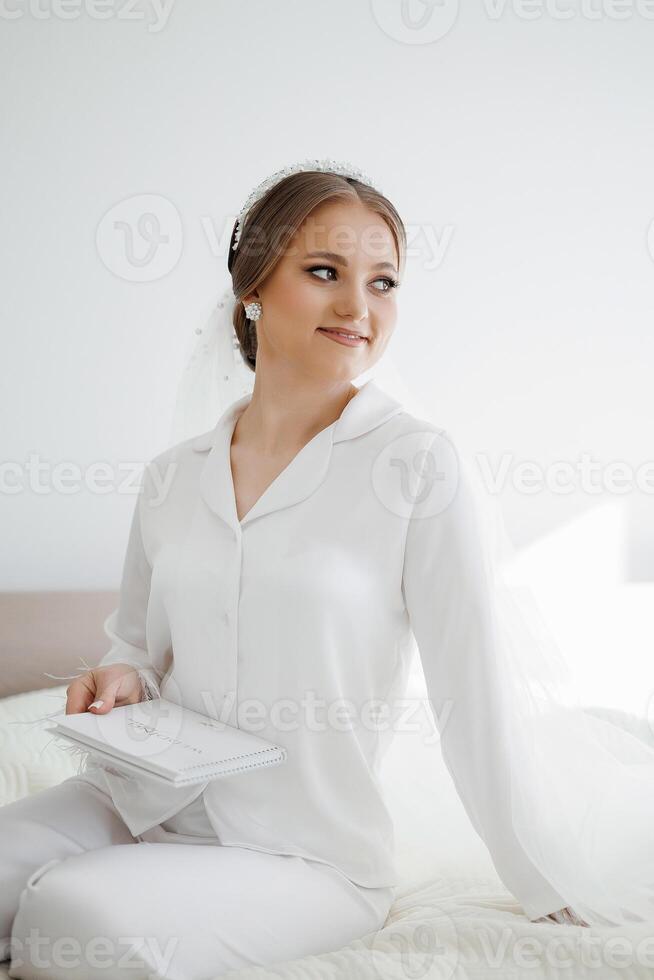 A woman in a white dress is sitting on a bed with a white sheet. She is holding a book in her lap photo