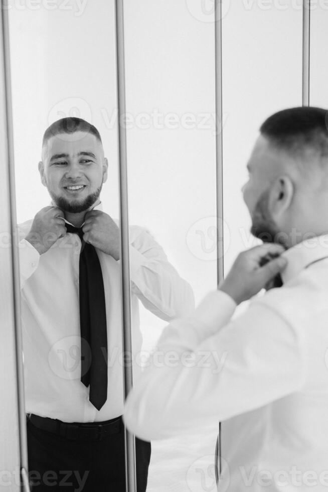 A man is getting ready for a formal event, putting on a black tie and adjusting it in the mirror photo