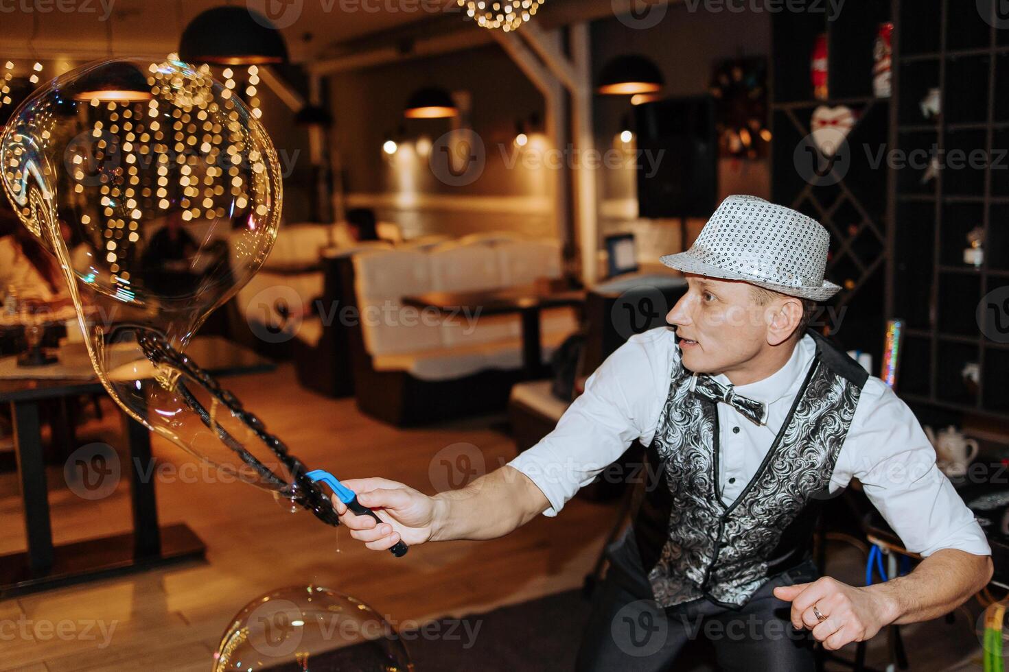 A man in a hat and vest is blowing bubbles. The bubbles are floating in the air and the man is holding a wand. The scene is lively and fun, with the bubbles adding a playful touch to the atmosphere photo