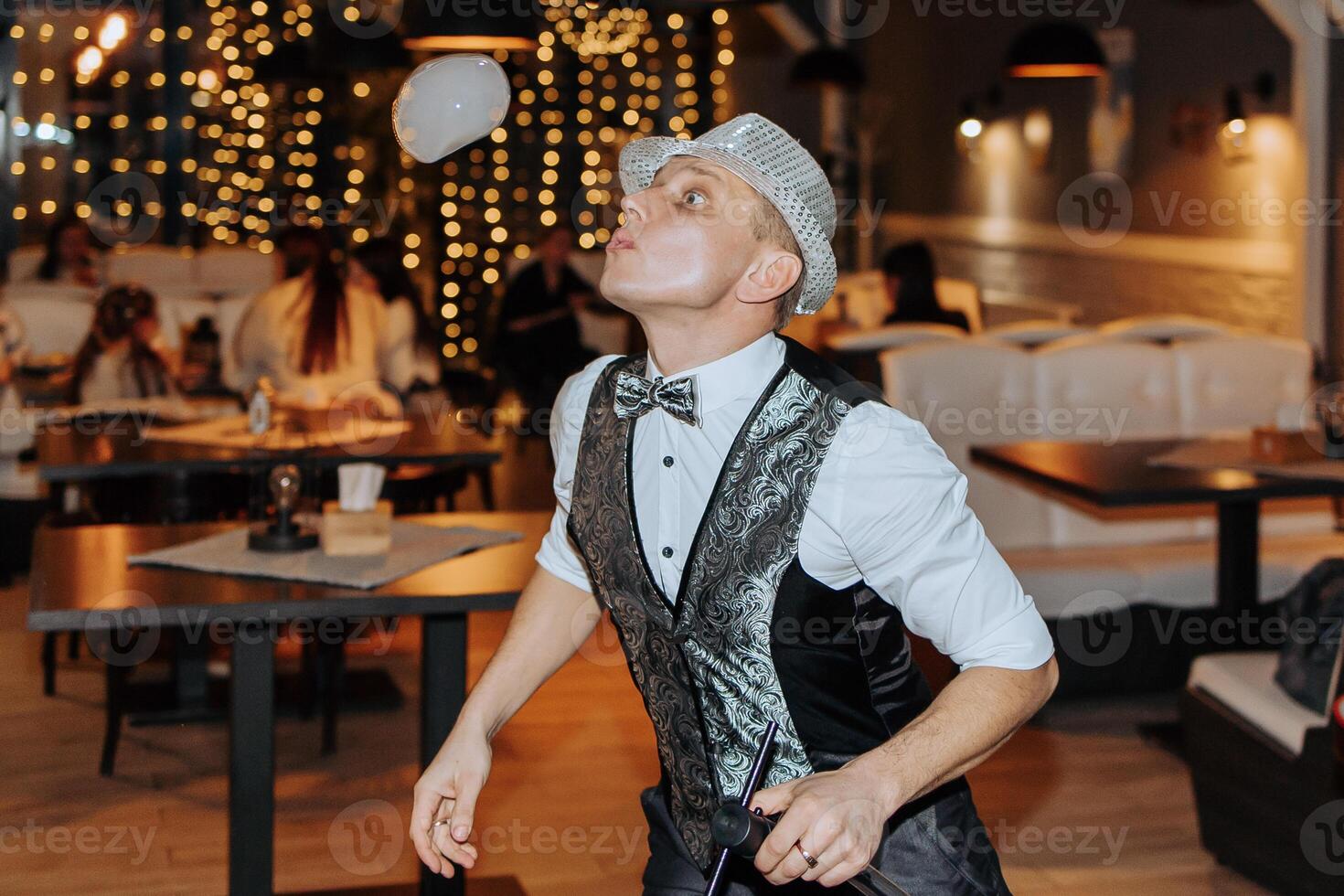 A man in a black vest and hat is juggling a ball. The scene takes place in a restaurant with tables and chairs photo