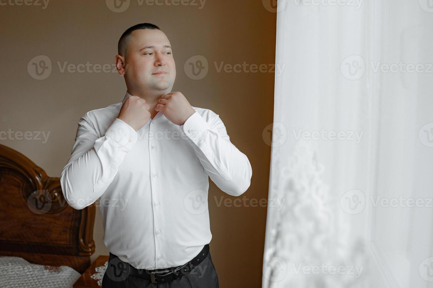 A man in a white shirt is getting dressed in front of a window. Scene is one of preparation and focus, as the man is carefully adjusting his collar and shirt photo