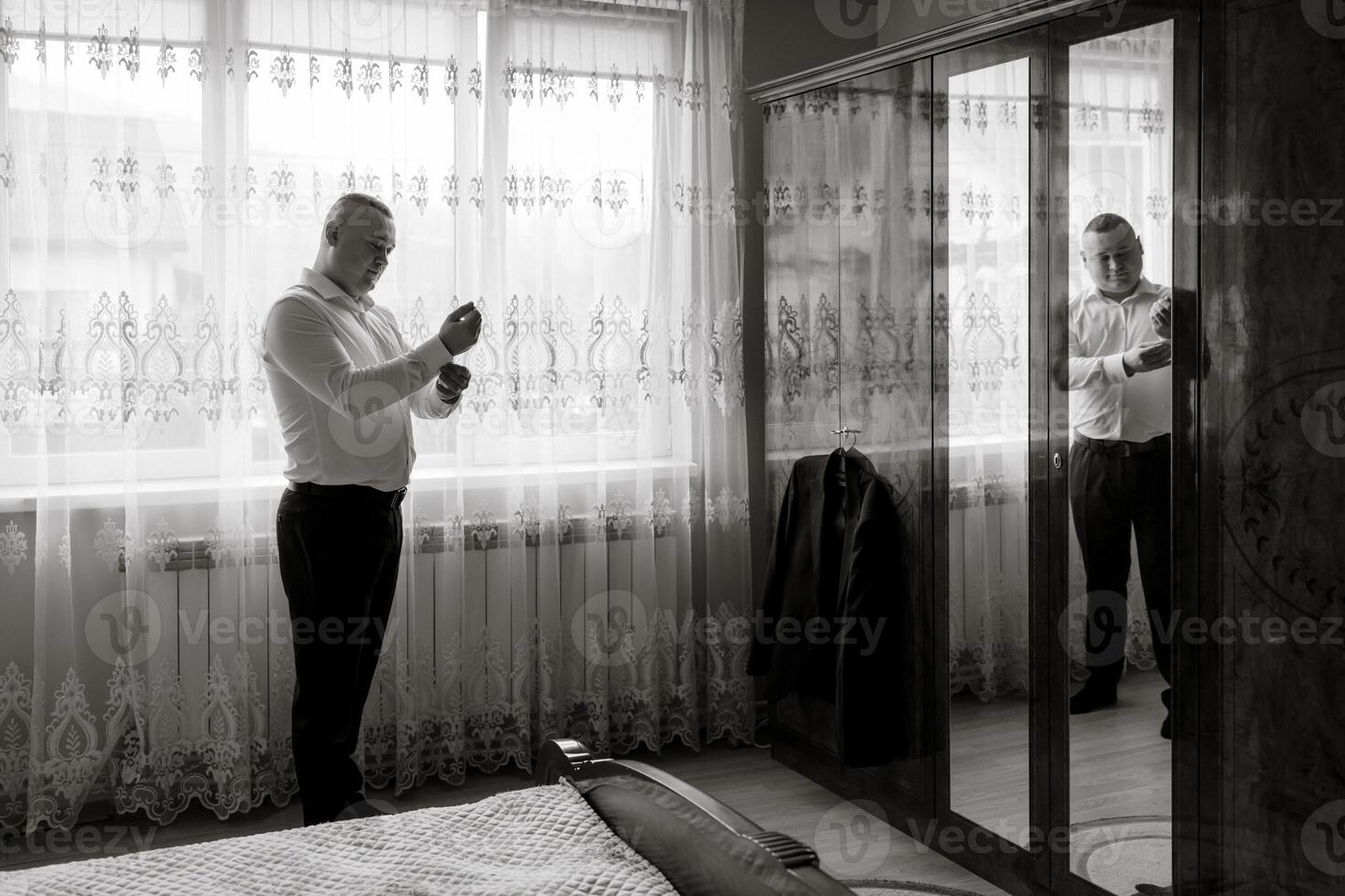 A man is getting ready for a formal event, putting on his suit and tie. He is standing in front of a mirror, adjusting his tie and looking at himself. The room is decorated with curtains and a bed photo