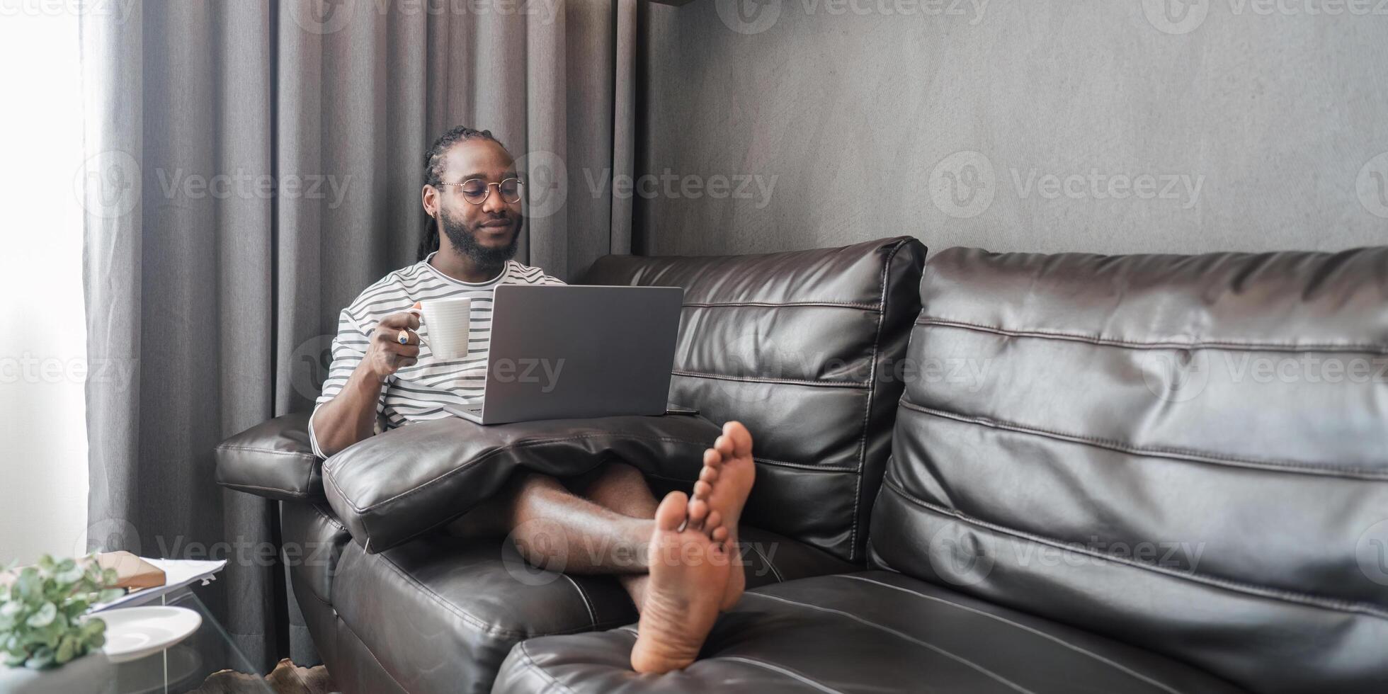 Young African American Man Relaxing on Couch with Laptop and Coffee in Modern Living Room photo