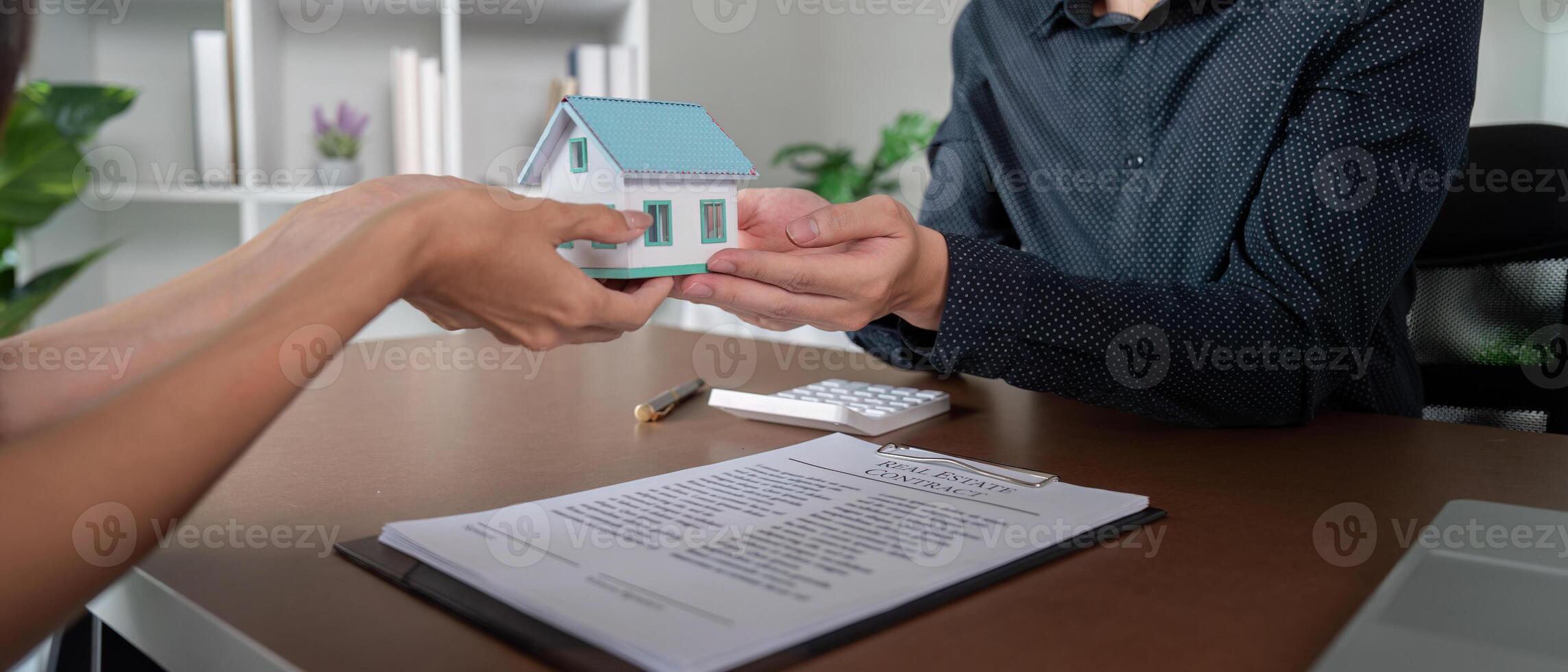 Real Estate Agent Handing Over House Model to Client with Contract and Calculator on Desk photo