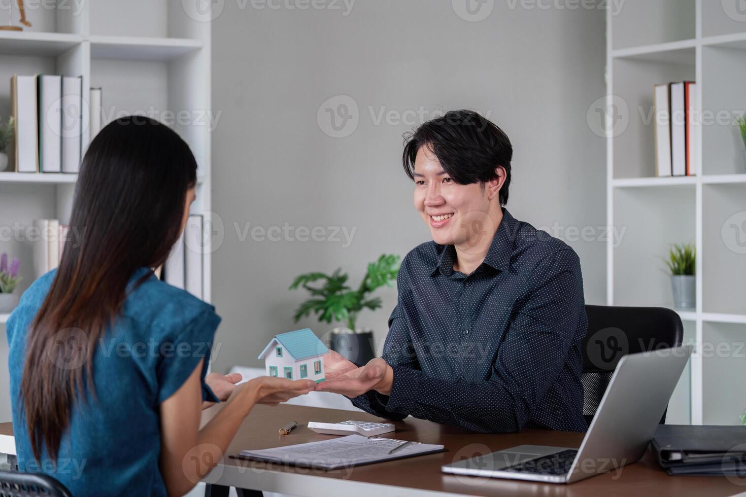 Real Estate Agent Showing Model House to Client in Modern Office Setting with Laptop and Documents photo