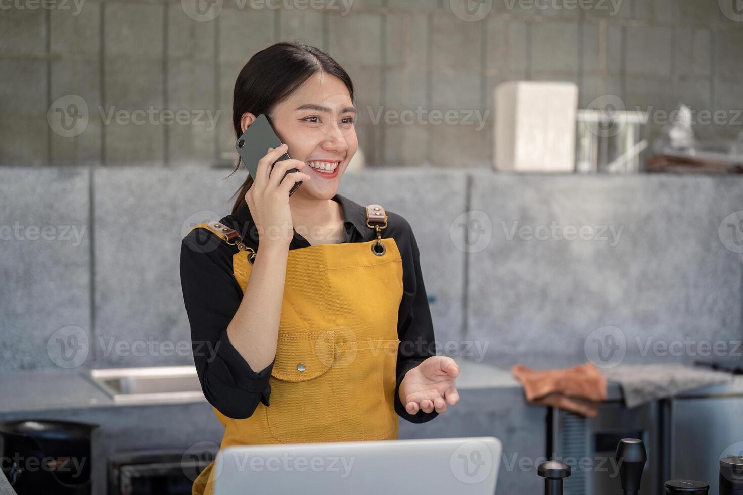 Young Barista in Yellow Apron Smiling While Talking on Smartphone in Modern Kitchen photo