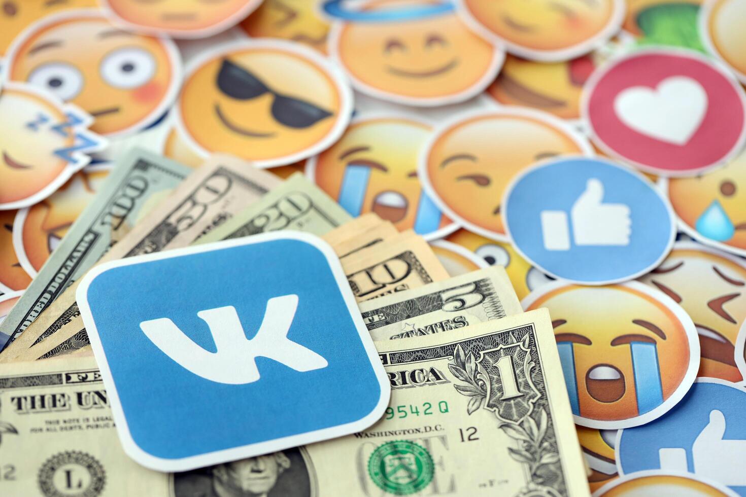 KYIV, UKRAINE - APRIL 20, 2024 VK logo and many usd money bills. Symbol of famous social network platform and dollar currency banknotes on lot of emoji reactions photo
